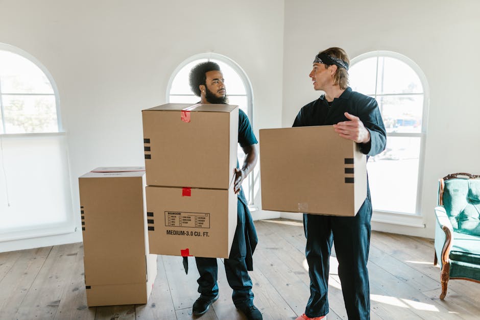 A professional mover from company name, dressed in dark clothing, is seen inside a residential property during a home relocation process, carefully lifting a green velvet sofa with button-tufted uphol