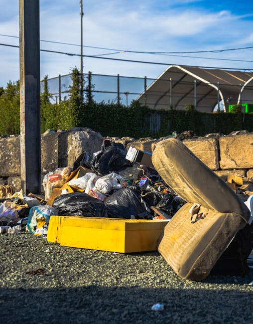 An outdoor scene showing a pile of bulky waste and discarded household items on a gravel surface adjacent to a stone wall, with black garbage bags, cardboard boxes, and a yellow plastic container amon