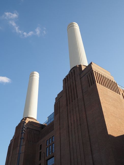 A view of the River Thames with a large industrial building featuring three tall chimneys on the opposite bank. In the foreground, part of a modern building's understructure is visible, supported by m